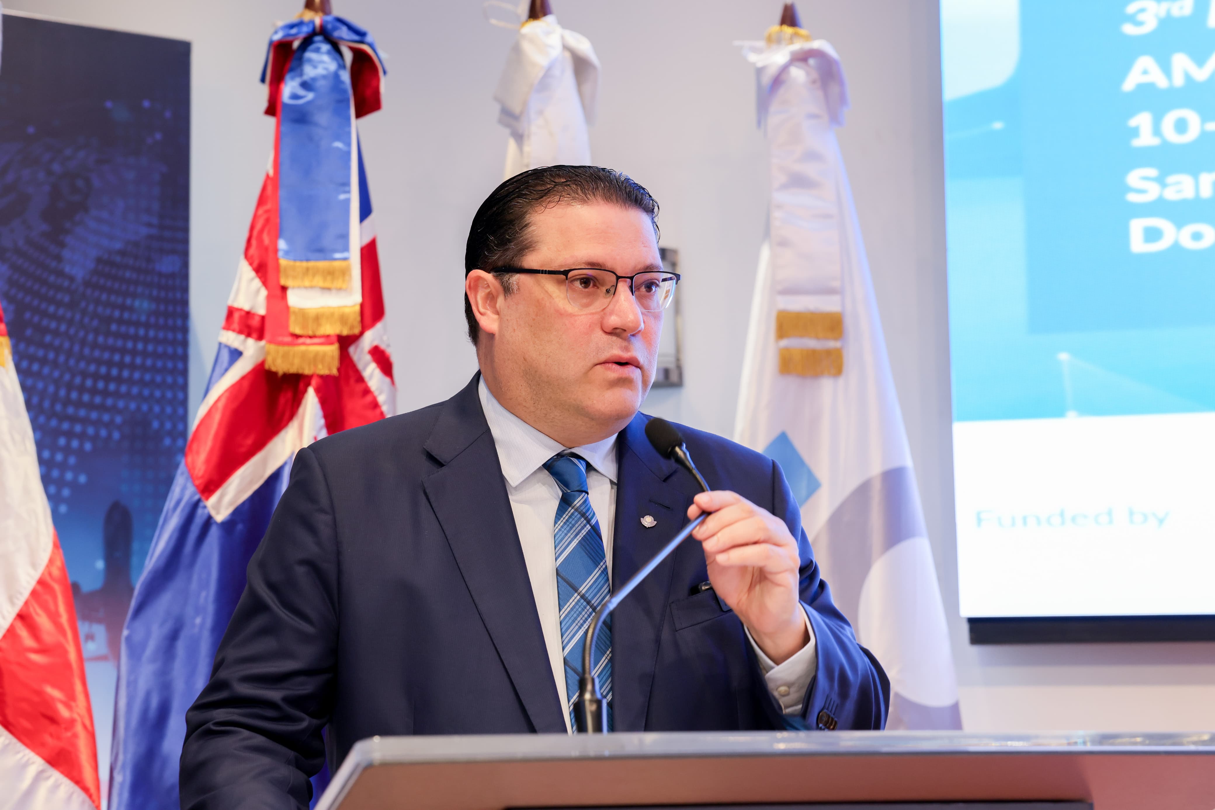Yayo Sanz Lovatón Titular De Aduanas parado frente a un podio, sosteniendo el micrófono, detrás de él se ven varias banderas incluidas la bandera dominicana y la de la DGA