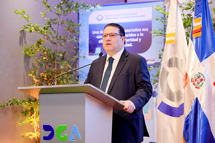 Yayo Sanz Lovatón Titular De Aduanas parado frente a un podio en la DGA, detrás de él se ven varias banderas incluidas la bandera dominicana y la de la DGA
