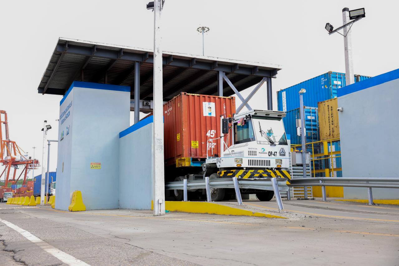 Vehículo de carga transportando un contenedor rojo a través de un punto de control en un puerto. La estructura tiene cámaras y equipos de inspección, rodeada por contenedores de distintos colores. La escena representa un proceso de revisión o escaneo de mercancías en una zona aduanera